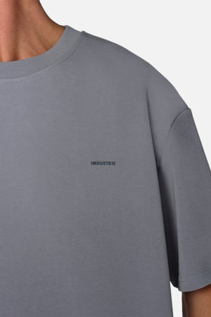 A person wearing an oversized fit Industrie The Newport Tee, made from 100% cotton, showcases the word "INDUSTRIE" printed in small letters on the chest against an off-white background.