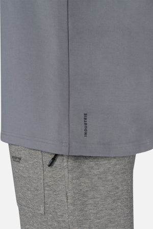 A close-up captures a person donning an oversized Industrie The Newport Tee in gray, paired with lighter gray pants. The shirt, made entirely from cotton, showcases "INDUSTRIE" near the hemline. The pants include a pocket accented by a black zipper. The backdrop is simple and light-hued.