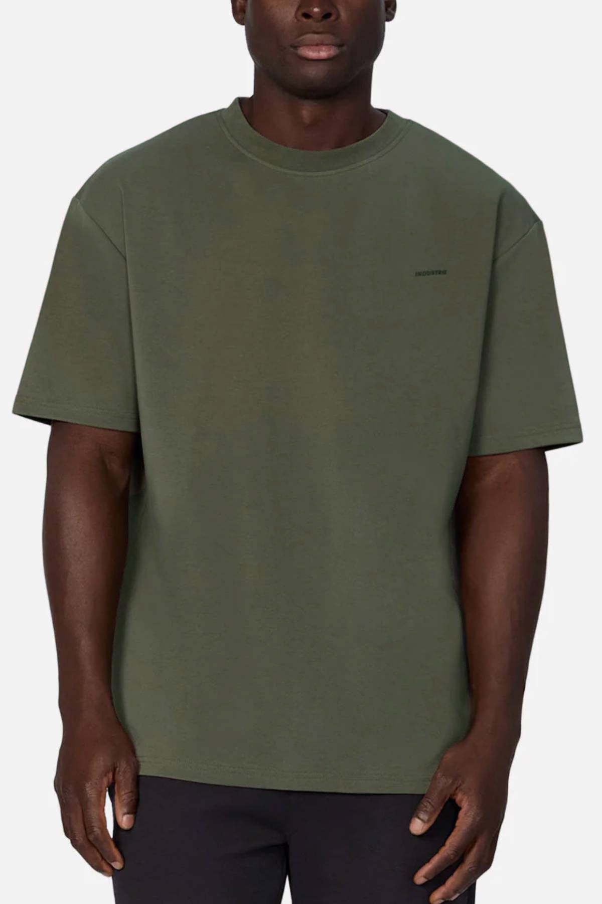 A man wearing an oversized olive green Industrie The Newport Tee, crafted from 100% cotton with subtle left chest text, stands against a plain light background.