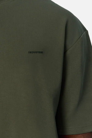Close-up of a person wearing the olive green Industrie The Newport Tee, featuring "INDUSTRIE" in small dark letters on the left chest. This 100% cotton tee has an oversized fit and is set against a plain light background.