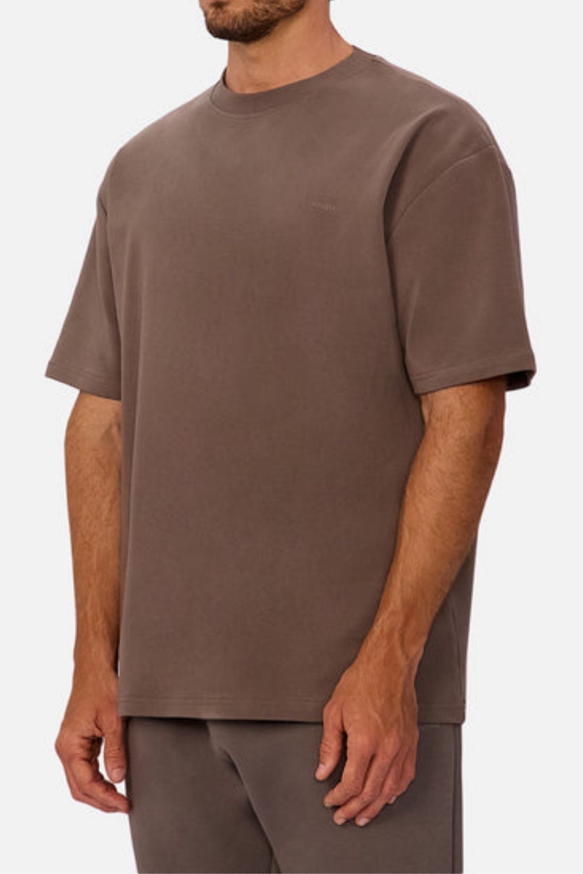 A man stands against a white background wearing Industrie The Newport Tee in brown, an oversized, short-sleeve 100% cotton shirt with a simple design and subtle chest logo, paired with matching pants.