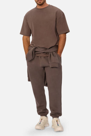 A man wearing the Industrie The Newport Tee, an oversized 100% cotton brown t-shirt, and matching sweatpants with a sweatshirt tied at his waist and white sneakers stands against a plain white background.