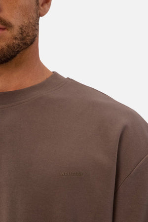 Close-up of a man wearing the Industrie The Newport Tee, an oversized brown crew-neck sweatshirt with "INDUSTRIE" subtly embossed on the chest. Only part of his face, neck, shoulder, and upper chest appear against a plain background.
