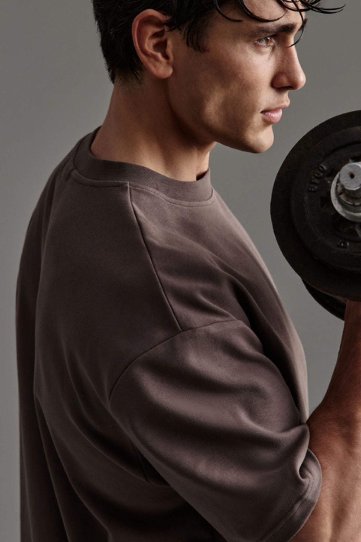 A man in an oversized Industrie The Newport Tee holds a dumbbell in one hand, gazing forward with a focused expression against a neutral background.