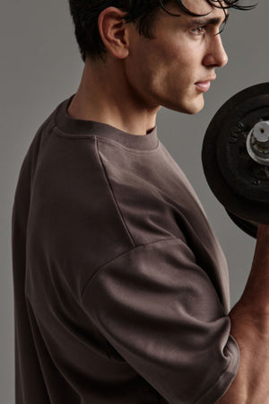 A man in an oversized Industrie The Newport Tee holds a dumbbell in one hand, gazing forward with a focused expression against a neutral background.
