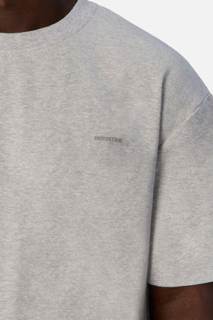 Close-up of a person wearing "Industrie The Newport Tee" in light gray, featuring the word "INDUSTRIE" subtly printed on the chest. Made from 100% cotton, the top half of the T-shirt and part of the sleeve reveal its oversized fit against a plain background.