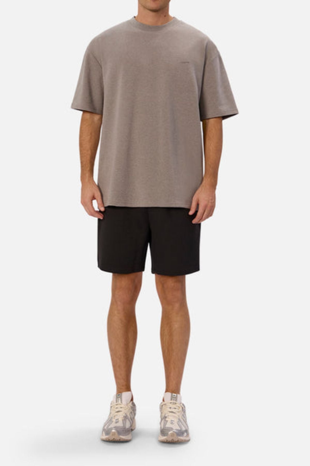 A person stands against a plain white background, wearing an oversized fit grey t-shirt, likely the Industrie The Newport Tee made from 100% cotton, paired with black shorts and beige sneakers.