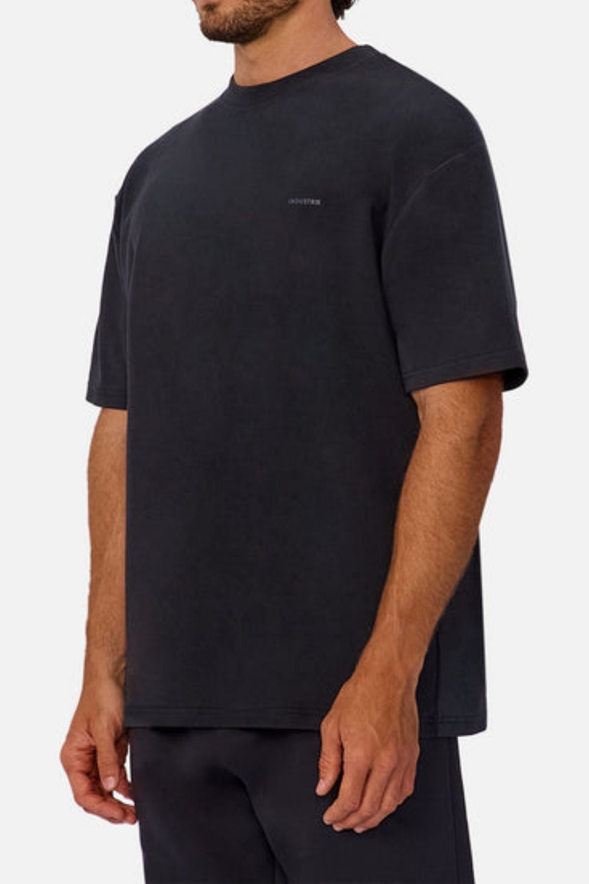 A man wears an oversized, short-sleeved black Industrie The Newport Tee with subtle chest text, paired with black pants, against a plain light background.