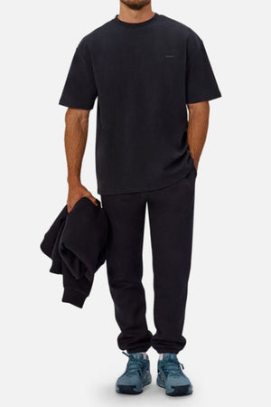 Wearing an Industrie The Newport Tee crafted from heavyweight fabric with black sweatpants and blue sneakers, a person stands against a plain light background, holding a folded black sweatshirt in one hand.
