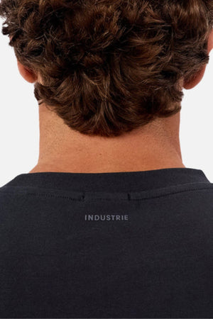 The image shows the back of a person's neck and head with curly brown hair, wearing an oversized Industrie The Newport Tee featuring "INDUSTRIE" printed near the collar on heavyweight fabric.