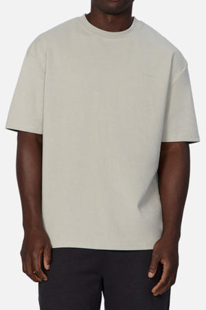 The individual dons a light gray Industrie The Newport Tee with an oversized fit, made from heavyweight fabric, paired with black pants against a white background. The round neckline enhances the relaxed mood, yet their head is not shown.