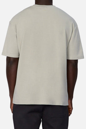 A person stands with their back to the camera, wearing Industrie's The Newport Tee in light gray and dark pants against a gray backdrop. The oversized tee is made from heavyweight fabric, showcasing the stylish back view of this casual outfit.
