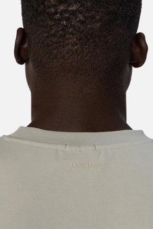 The image shows the back of a person wearing an oversized light-colored T-shirt labeled "INDUSTRIE" below the neckline, featuring Industrie’s The Newport Tee in heavyweight fabric for a stylish and comfortable look.