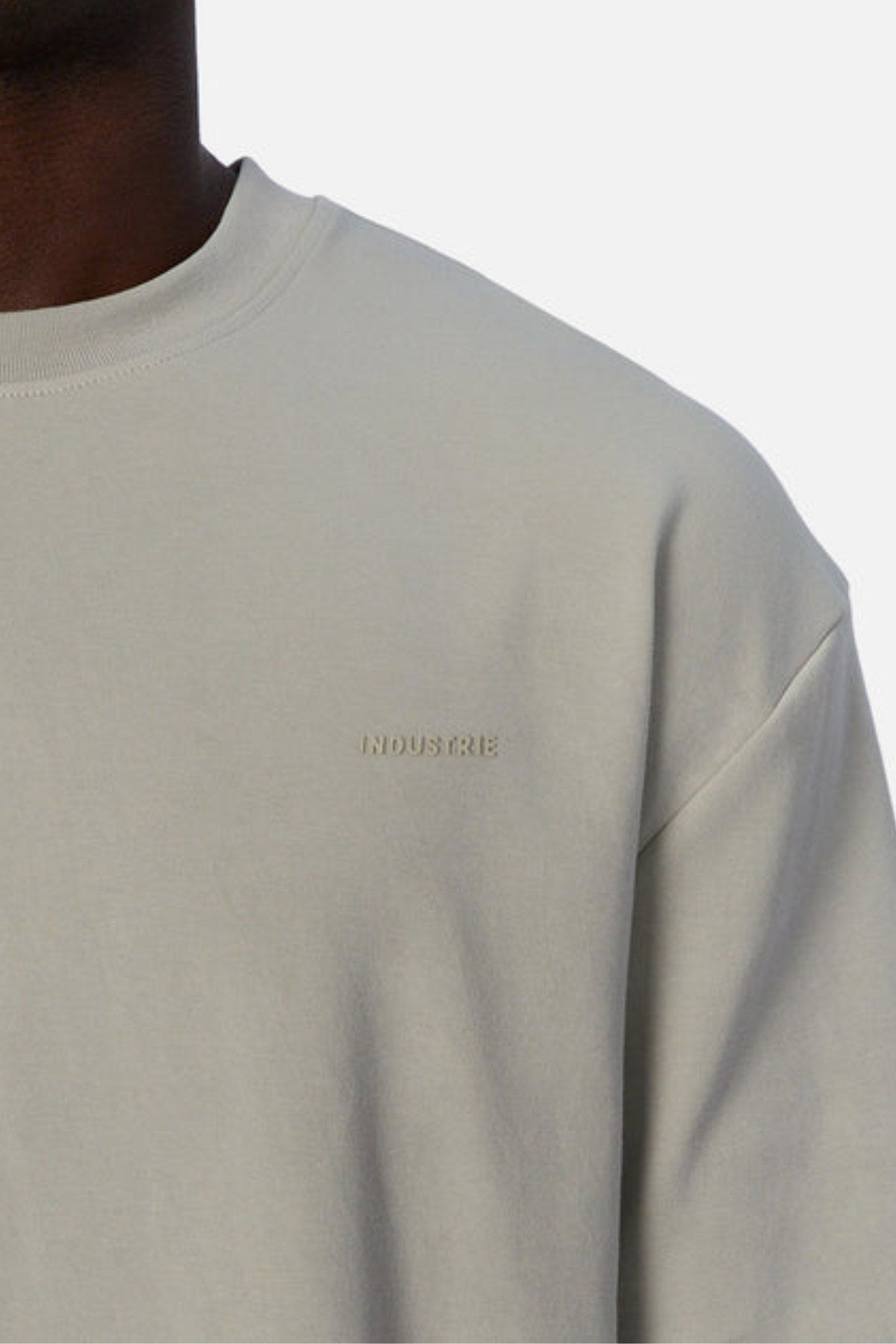 Close-up of someone in "Industrie The Newport Tee," an oversized fit light gray sweatshirt with subtle "INDUSTRIE" chest embroidery. The heavyweight fabric exudes durability and comfort, all captured against a plain white background.
