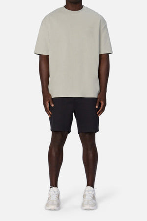 A person stands against a plain backdrop in Industrie's The Newport Tee in light gray, black shorts, and white sneakers. The oversized fit and heavyweight fabric of the tee enhance the casual style. Their arms rest relaxed at their sides.