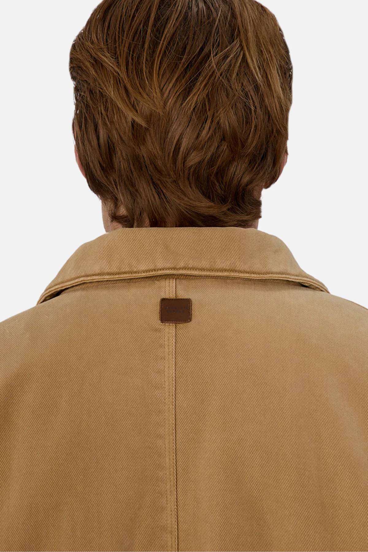 A person with light brown, wavy hair is shown from behind, wearing the Industrie The Oklahoma Jacket in tan, featuring a visible seam and a small brown square patch near the collar. The background is plain and light-colored.