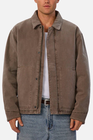Someone stands facing forward against a plain light background, wearing Industrie The Oklahoma Jacket—a loose-fitting brown jacket with a collar, button closure, and two front pockets—layered over a white shirt and blue jeans.