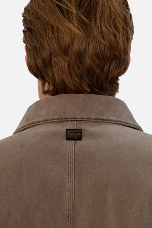 A person with light brown hair is shown from behind, wearing the Industrie The Oklahoma Jacket in brown, featuring a small black and orange INDUSTRIE label on the upper back, showcasing its versatile outerwear style.