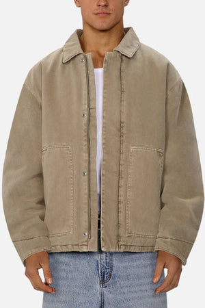 Wearing the Industrie The Oklahoma Jacket in tan over a white shirt and blue jeans, a person stands against a plain light background. This casual-fit jacket features a collar, metal snap buttons, front pockets, and functional design details.