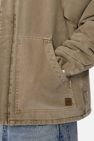 A person wearing the Industrie The Oklahoma Jacket in tan, featuring a practical design and large front pocket, has their hand partially inside the pocket and is also dressed in light blue jeans.