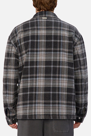 A person facing away from the camera wears the Industrie The Phoenix Jacket, a gray and black plaid heavyweight shirt jacket with long sleeves, paired with dark gray pants.