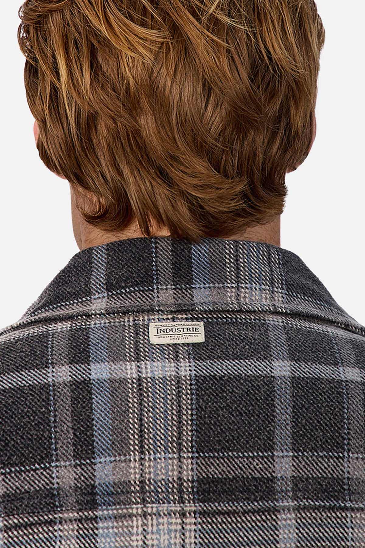 A person with light brown hair is seen from behind wearing the Industrie The Phoenix Jacket, a heavyweight plaid shirt jacket in grey, black, and blue shades. A small white "INDUSTRIE" label is sewn onto its upper back.