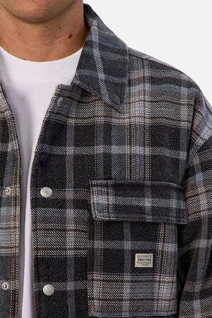 A person wears the Industrie The Phoenix Jacket—a heavyweight black, gray, and blue plaid shirt jacket—over a white t-shirt. A small rectangular "Industrie" label is visible on the front pocket; only the lower half of their face is shown.