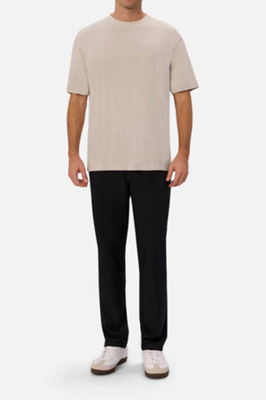 A person wearing a beige T-shirt, Industrie The Tech Pinehurst Chino Pant, and white sneakers stands against a plain white background. The casual outfit, featuring recycled polyester for added flexibility, is minimalistic yet sophisticated.