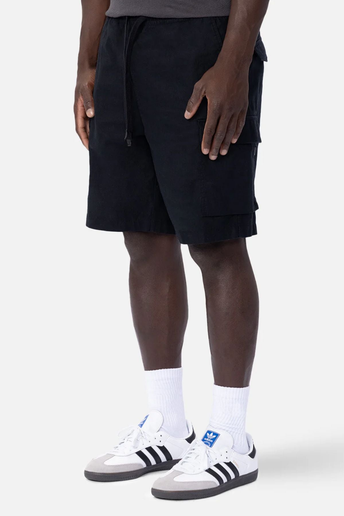 A person wears Industrie The Platoon Combat Short, a dark t-shirt, white crew socks, and white Adidas sneakers with black stripes, standing against a plain light background.