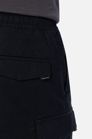 Close-up of a person wearing Industrie The Platoon Combat Short in black, featuring an elastic waistband, flap pockets with a small tag, paired with a tucked-in dark gray shirt.