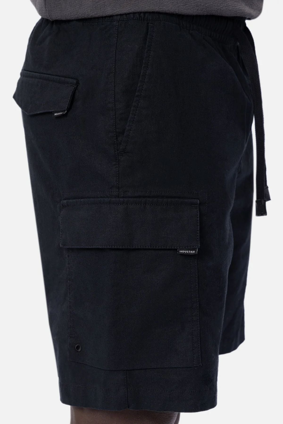 A close-up side view of a person wearing Industrie The Platoon Combat Short in black, featuring a four-pocket design with large flap pockets and an elastic waistband, set against a plain light-colored background.