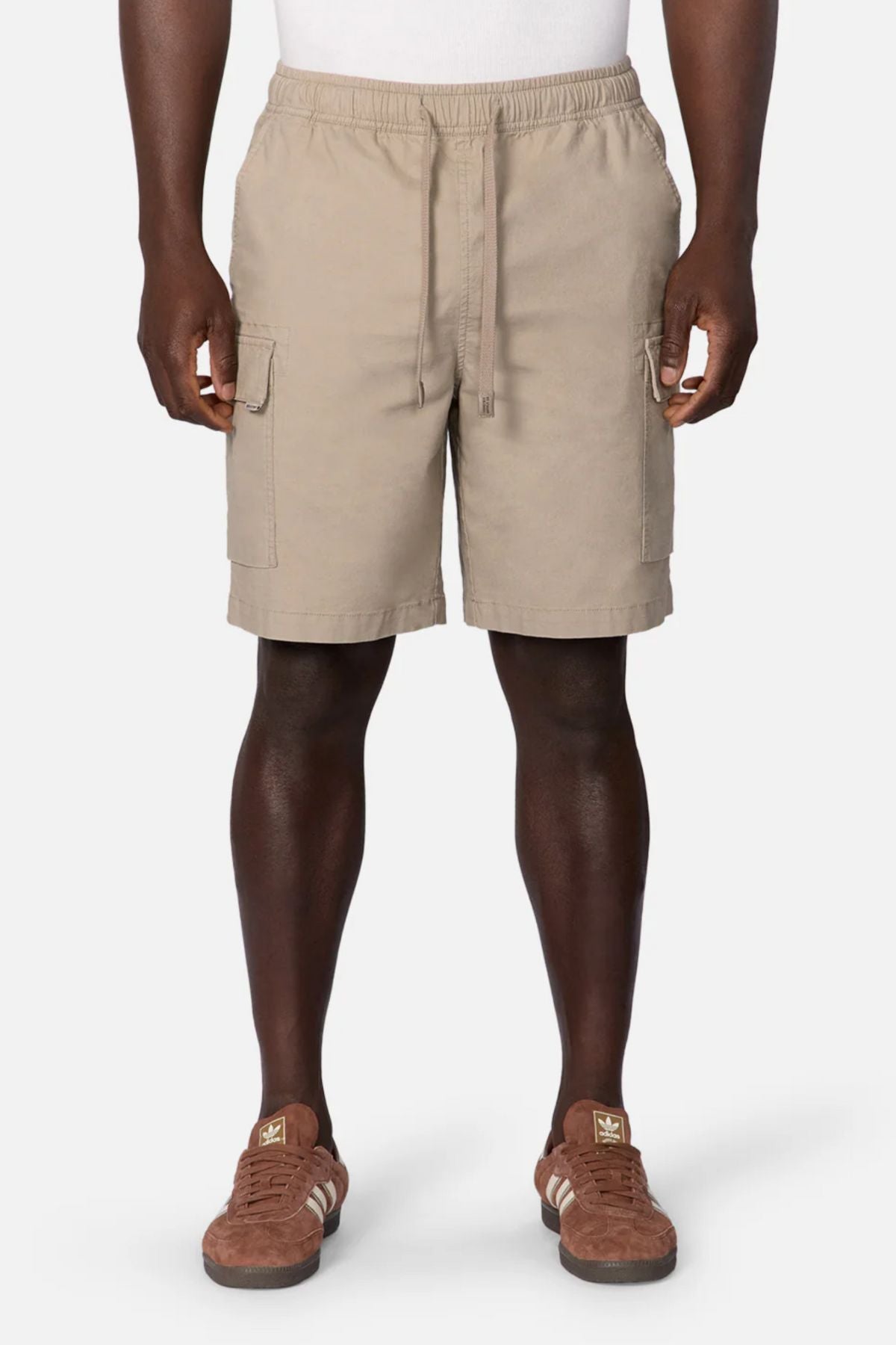 A person wearing Industrie The Platoon Combat Short in beige, featuring an elastic waistband and four pockets, with a white shirt and brown Adidas sneakers, stands against a plain white background.