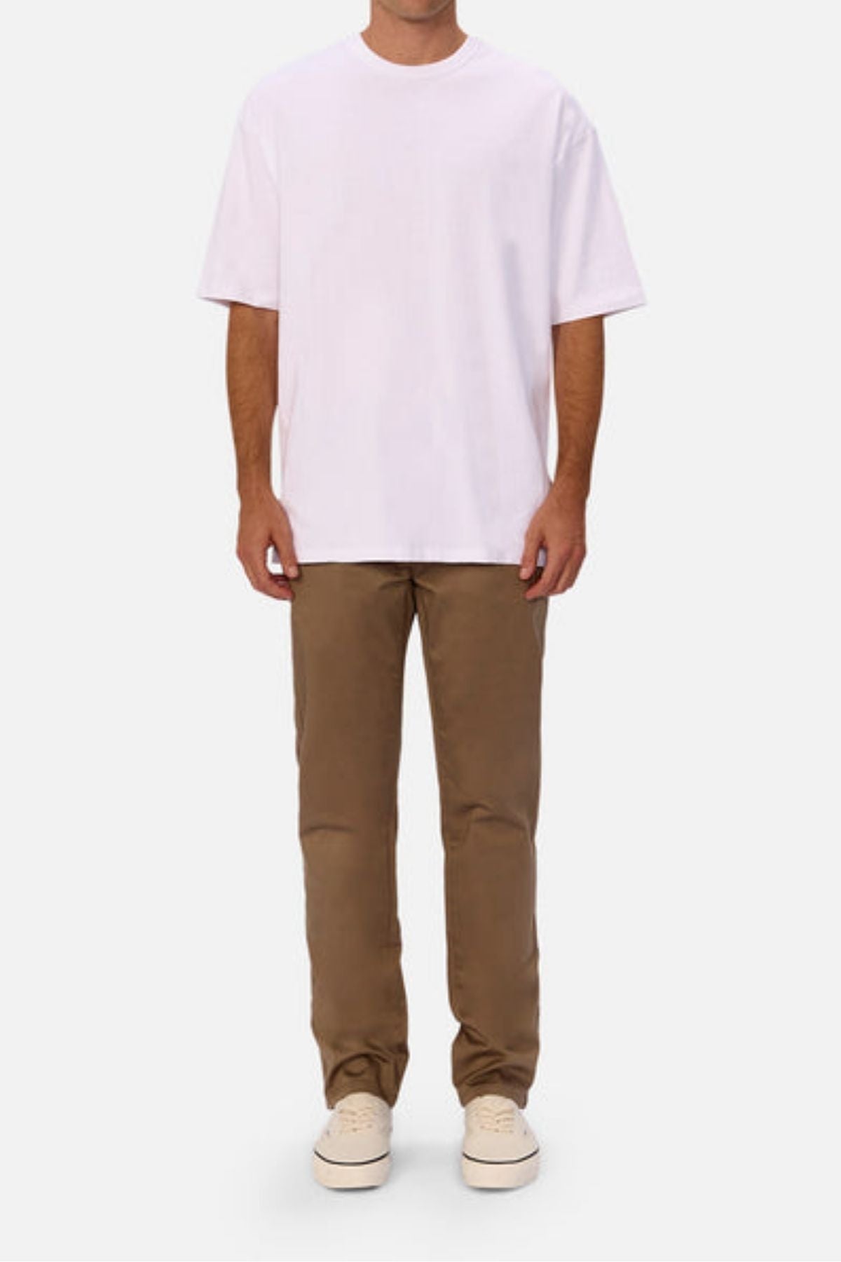 A person stands against a plain background wearing a loose-fitting white T-shirt, Industrie The Regular Cuba Chino Pant in tan with a wider leg fit, and light-colored sneakers. The person's head is not visible in the image.