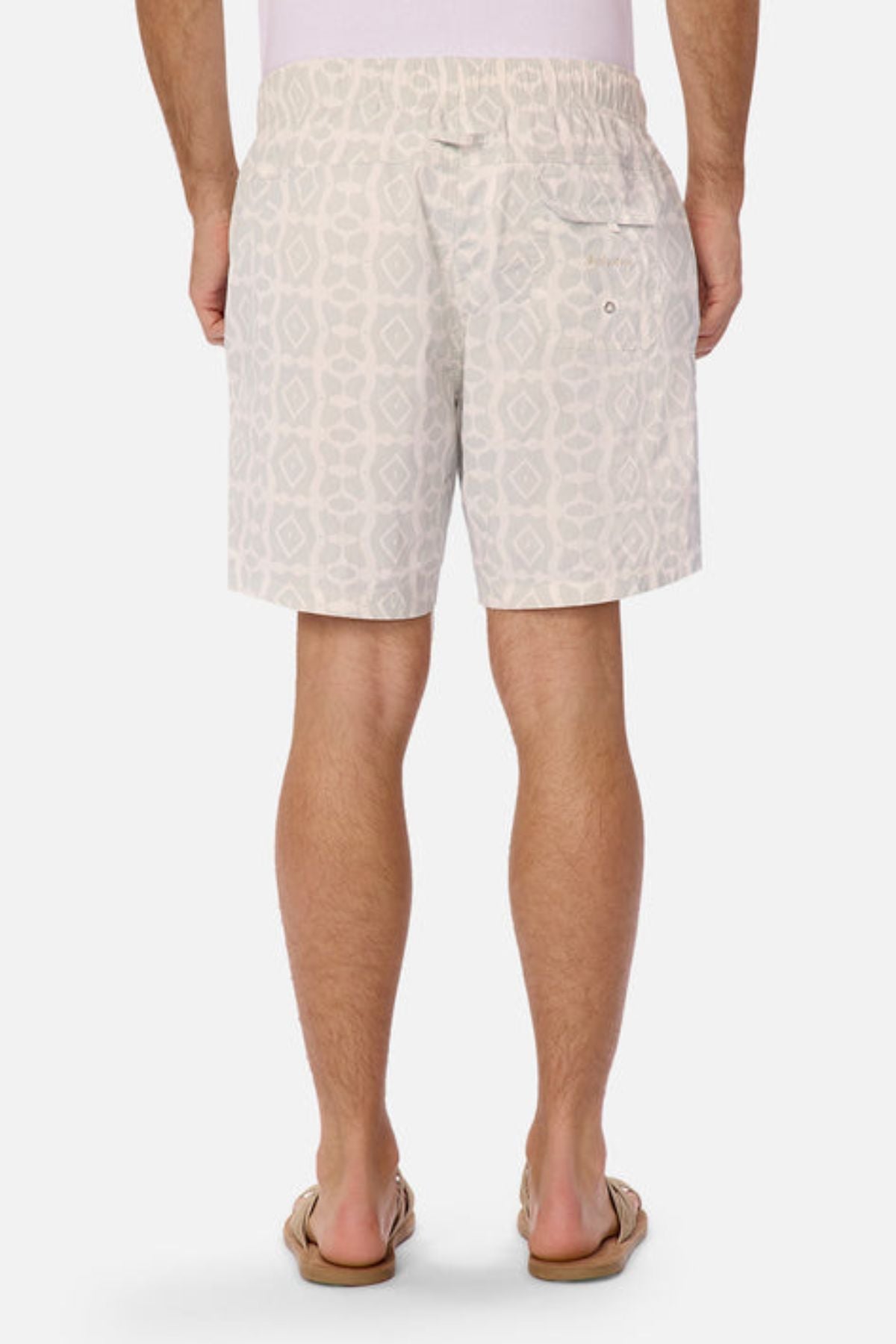 From the back, a person is wearing Industrie The Saratoga Swim Short in a distinctive light gray print. These shorts come with an elastic waistband, a right-side pocket, and are designed to be quick-drying. Beige flip-flops complete the ensemble.