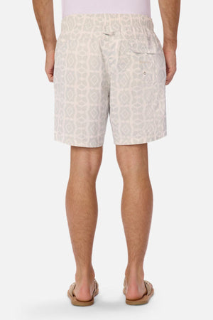 From the back, a person is wearing Industrie The Saratoga Swim Short in a distinctive light gray print. These shorts come with an elastic waistband, a right-side pocket, and are designed to be quick-drying. Beige flip-flops complete the ensemble.