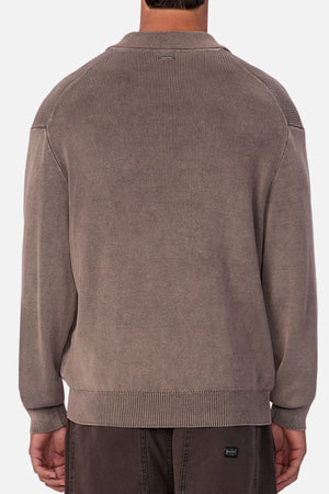 A person is seen from behind in a loose, long-sleeve taupe Industrie The Sawtelle Knit sweater made of 100% cotton with ribbed cuffs and hem, paired with dark pants against a plain neutral background.