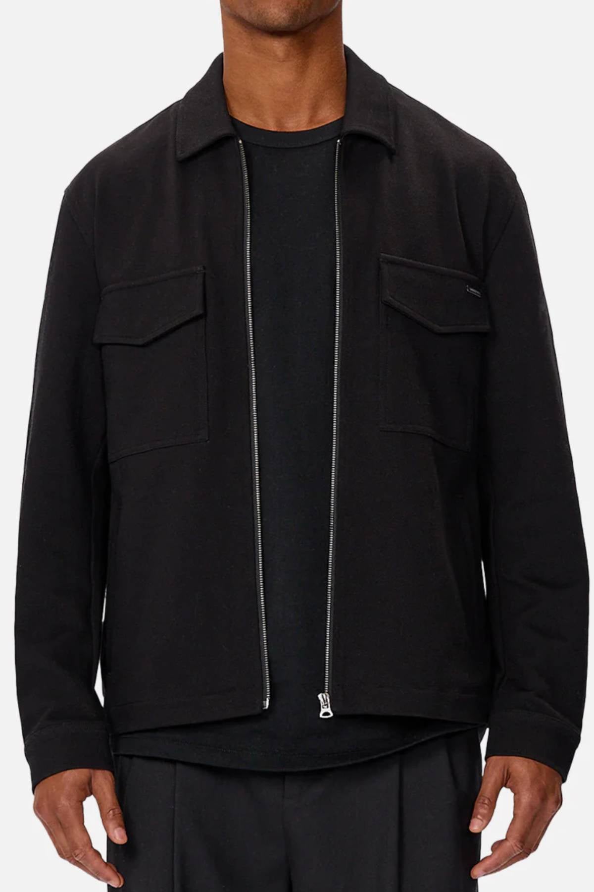 A person is shown from shoulders to hips wearing a black Industrie The Southport Jacket with a front zip and two chest pockets over a black shirt, set against a plain light background.