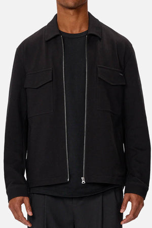 A person is shown from shoulders to hips wearing a black Industrie The Southport Jacket with a front zip and two chest pockets over a black shirt, set against a plain light background.