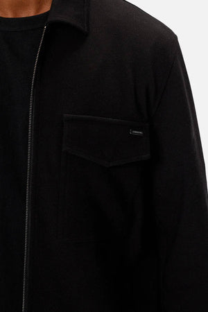 A close-up of a person wearing the Industrie The Southport Jacket in black, featuring a front zip and flap chest pocket over a black shirt. Only the upper torso and part of the neck are visible against a plain light background.