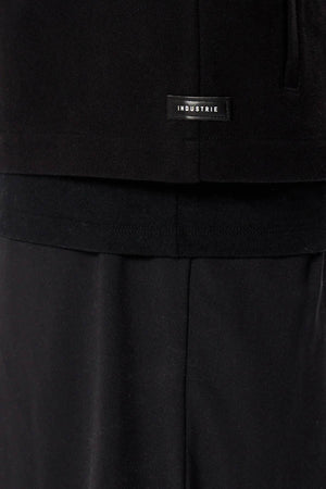 Close-up of a person in black pants and a black shirt layered over another black top, featuring the Industrie logo—a subtle nod to the minimalist design of Industrie The Southport Jacket.