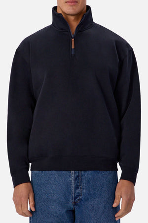 A person in an Industrie The Staunton Track Top—a black quarter-zip pullover sweater—and blue jeans stands against a plain light background.