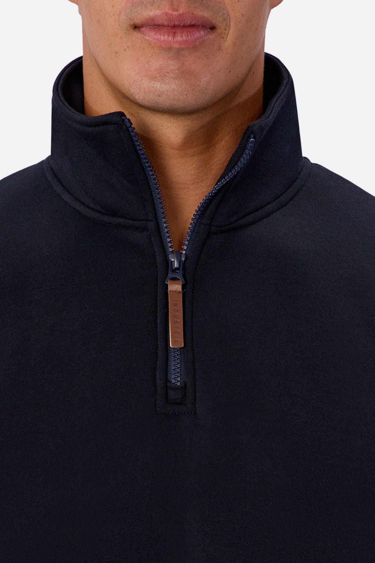 A close-up shows a person wearing the Industrie The Staunton Track Top, a men's casual quarter-zip pullover with a stand-up collar and leather-accented pull tab. Only the lower face and upper torso are visible.