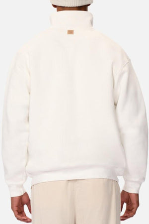 A person is seen from behind wearing the Industrie The Staunton Tracktop in white with a high collar, branded trims, and a brown tag below the collar. They pair it with beige pants and a cozy white knit beanie.