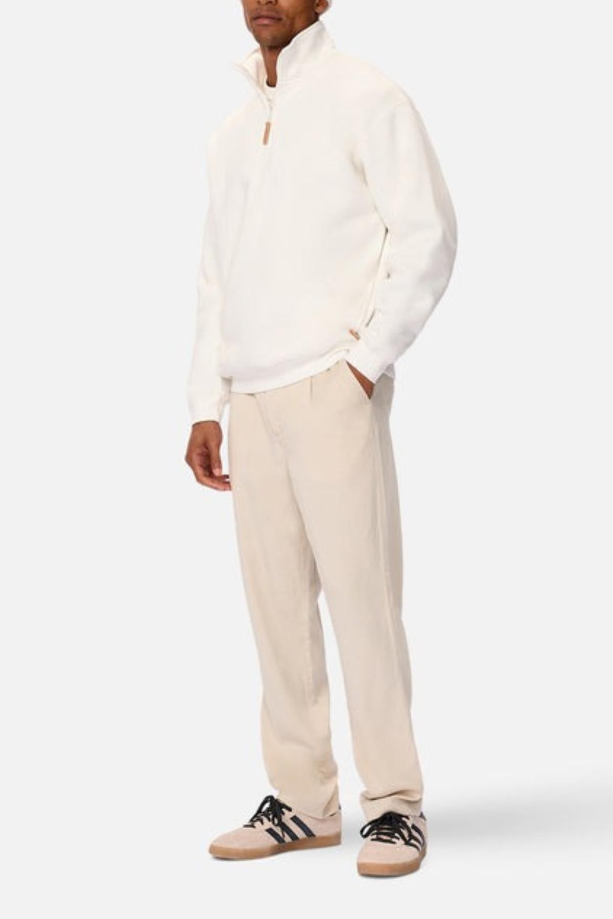 A man stands hands in pockets, wearing the Industrie The Staunton Tracktop with fleece lining, light beige pants, and beige sneakers with black laces against a plain light background.