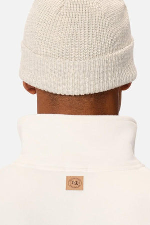 A person seen from behind wears a cream beanie and the Industrie The Staunton Tracktop, a white jacket with a high collar, branded trims, and a small brown logo patch at the back, set against a plain light background.