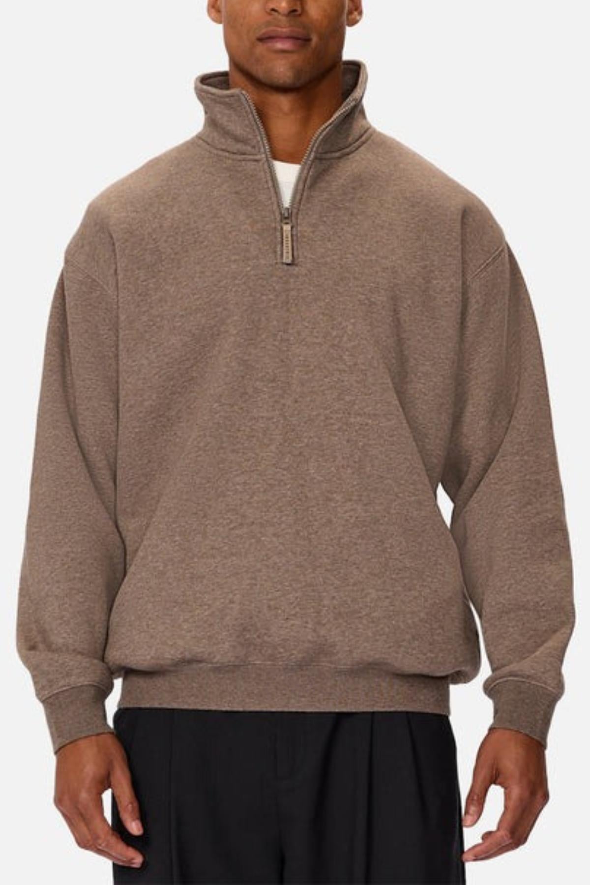 A man wearing the Industrie The Staunton Tracktop, a brown quarter-zip pullover with branded trims and a high collar, paired with black pants, stands against a plain light background.
