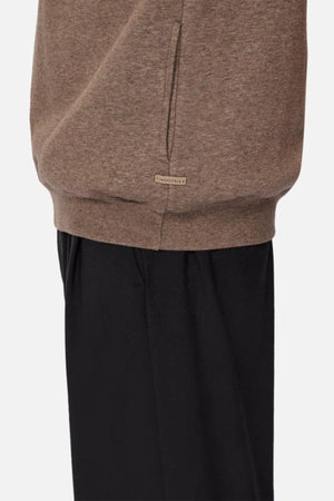 A person models Industrie The Staunton Tracktop in brown, featuring a small label near the hem, paired with loose black pants, shown from the waist down against a plain background.