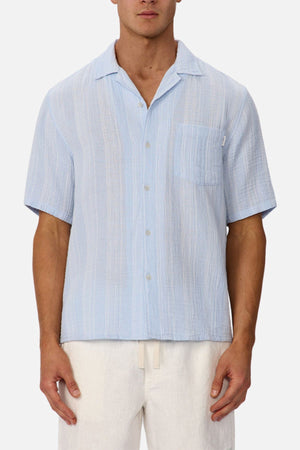 A man wearing the Industrie The Striped Seguro Short Sleeve Shirt, paired with white pants, stands against a plain light background.
