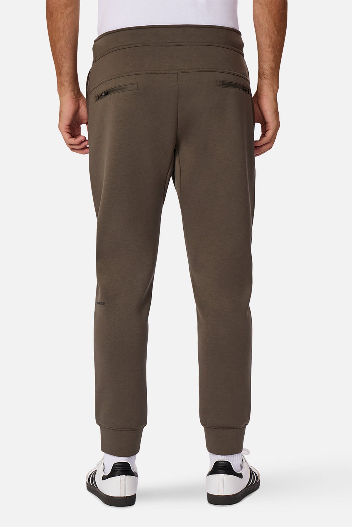 Someone wearing the Industrie The Tech Armoured Track Pant in olive green with zippered back and combat pockets, paired with white sneakers, stands facing away with hands by their sides against a plain white background.
