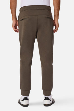 Someone wearing the Industrie The Tech Armoured Track Pant in olive green with zippered back and combat pockets, paired with white sneakers, stands facing away with hands by their sides against a plain white background.